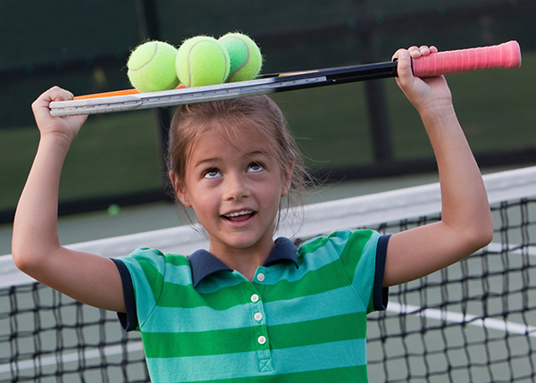 After her kids tennis lesson, a girl in a green striped polo goofs off with her tennis racquet.